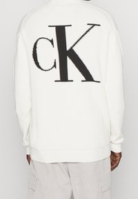 Weißer, gerippter Pullover mit großem schwarzem "ck"-Logo auf der Rückseite. Lange Ärmel mit lockerem Sitz und Rundhalsausschnitt.