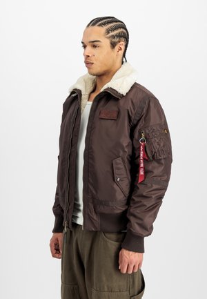 Chaqueta bomber marrón con cuello de borreguillo, cierre de cremallera, bolsillos laterales y un detalle de etiqueta roja en la manga. Textura acolchada en el cuerpo.