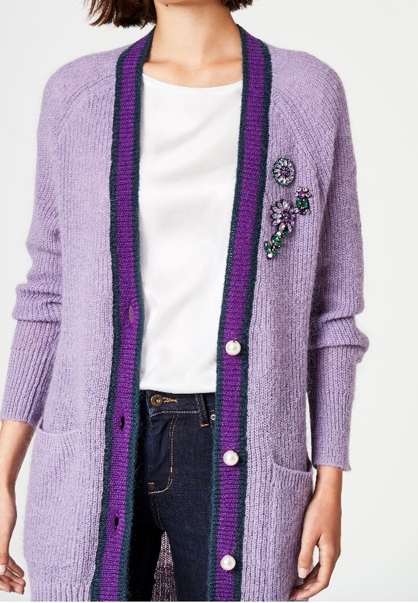 Ljuslila stickad cardigan med en mörkgrön och lila randig kant, prydd med pärlknappar och dekorativa blommor.