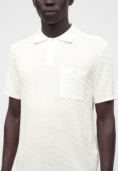 Homme portant un polo blanc texturé à manches courtes avec une poche poitrine boutonnée sur un fond uni.