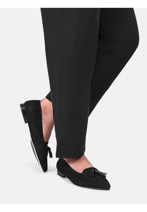 Voeten in zwarte suède loafers met puntige neus en kwastjes, gecombineerd met zwarte cropped broek tegen een effen achtergrond.