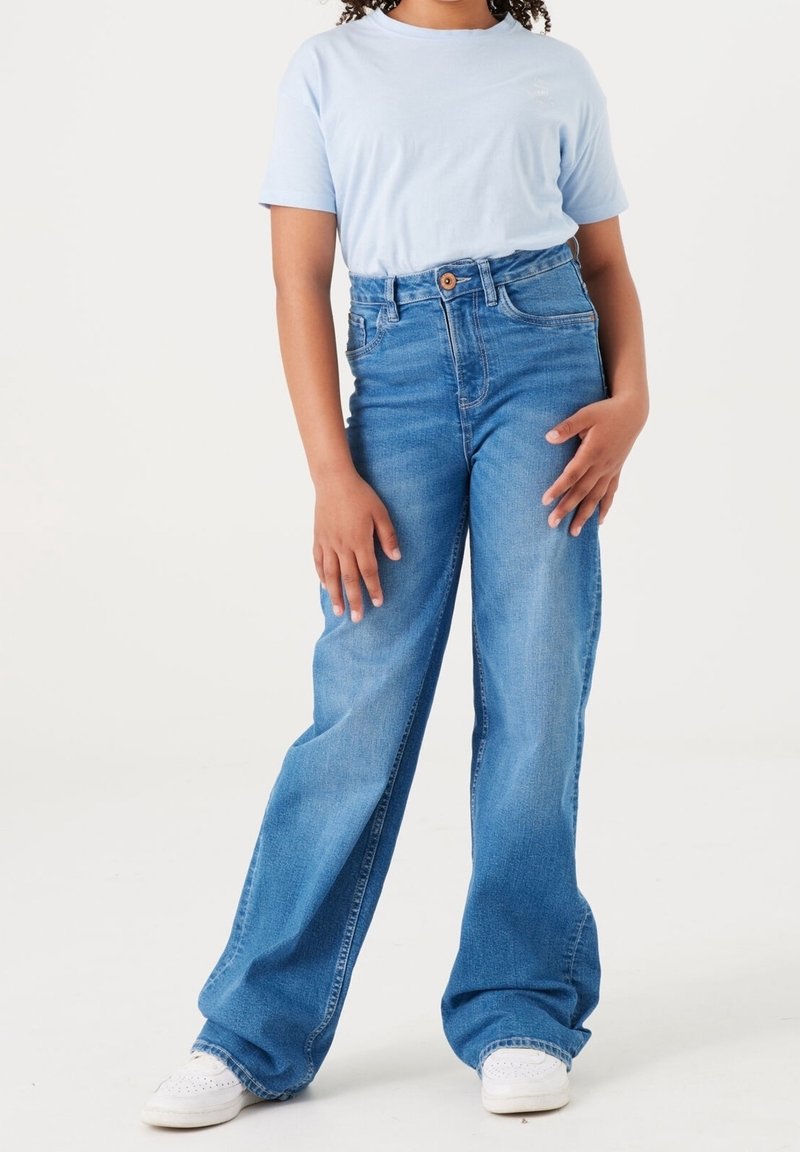 T-shirt bleu clair associé à un jean bleu à taille haute et à jambe large, avec une fermeture à bouton et une texture en denim classique. Des baskets blanches complètent le look.