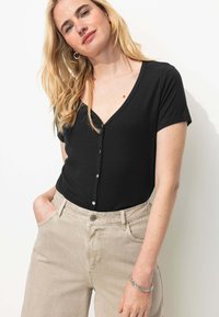 Haut noir ajusté à côtes avec de courtes manches et un décolleté en V profond, orné de boutons métalliques sur le devant. Associé à un pantalon beige clair.