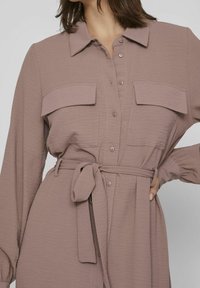 Dlouhé rukávy šaty v barvě mauve s předním zapínáním na knoflíky, dvěma náprsními kapsami, páskem v pase a texturovanou tkaninou.