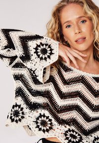 Häkelpullover mit Zickzack- und Blumenmustern in Schwarz und Creme. Mit weiten Glockenärmel und einem gewellten Dekolleté. Leichte Textur.