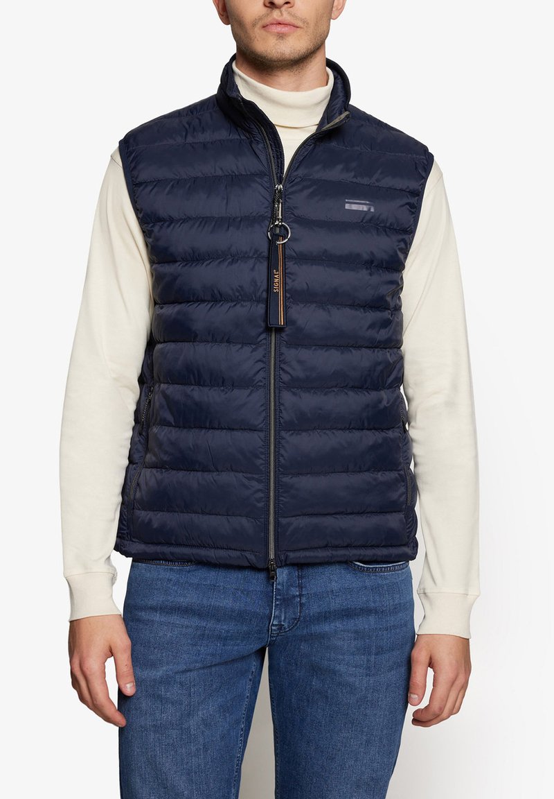Marineblå puffer vest med vertikal quiltet design, lynlåslukning og sidelommer. Båret over en cremefarvet turtleneck og kombineret med blå jeans.
