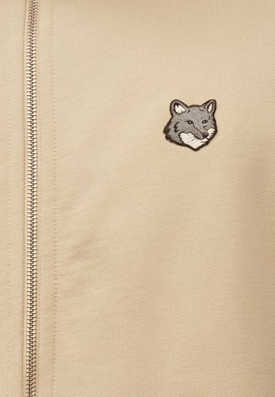 Μπεζ τζάκετ με φερμουάρ στο μπροστινό μέρος και μικρό κεντημένο γκρι κεφάλι αλεπούς στην αριστερή πλευρά του στήθους.