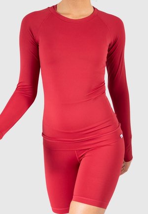 Woman wearing fitted long-sleeve red top and matching red cycling shorts against plain light background.