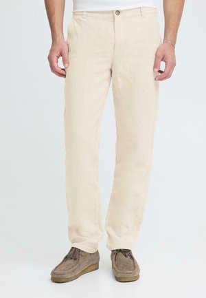 Beige Leinenhose mit geradem Bein. Verfügt über einen Knopfverschluss, Gürtelschlaufen und eine strukturierte Stoffoberfläche. Kombiniert mit hellbraunen Schuhen.