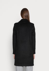 Melnā vilnas mētelī ar pielāgotu piegriezumu, garām piedurknēm un iegrieztu apkakli. Iekļautas pogu detaļas uz piedurknēm; gluda tekstūra, līdz pusei augšstilbam.