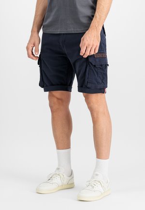 Jambes d’homme portant un short cargo bleu marine, des chaussettes blanches montantes et des baskets blanches, debout sur un fond clair uni.