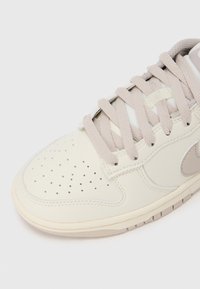 Tennisking, millel on sile kreemjas nahast pealiskate, beežid paelad, auguauguga varbakarp ja helehall Swooshi logo küljel. Pehme tekstuur.