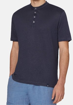 Mann trägt ein dunkel marineblaues Henley-Shirt mit kurzen Ärmeln und vier Knöpfen sowie hellblaue, strukturierte Shorts, Hände entspannt an den Seiten.