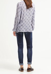 Kvinna som står vänd bort och bär en mönstrad långärmad blus, mörka uppvikta jeans och bruna oxfordskor på vit bakgrund.
