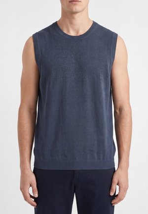 Uomo che indossa un gilet di maglia blu navy senza maniche e pantaloni scuri, in piedi davanti a uno sfondo bianco semplice.