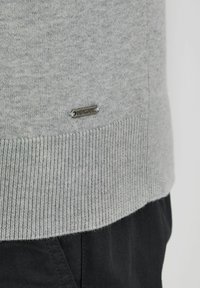 Suéter gris claro con textura de punto, que presenta un dobladillo acanalado. Incluye una pequeña etiqueta de metal con "INDICODE" en el lado inferior.