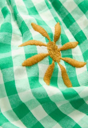 Grön och vit rutigt tyg med en framträdande broderad soldesign i guldtråd, med strukturella detaljer på solens strålar.