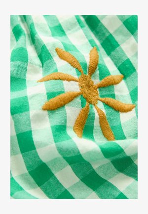 Grön och vit rutigt tyg med en framträdande broderad soldesign i guldtråd, med strukturella detaljer på solens strålar.