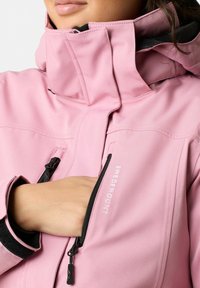 Giacca impermeabile rosa con colletto alto, tasche con cerniera e dettagli con logo. Presenta una texture liscia e polsini regolabili.