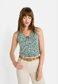 Top sin mangas verde con patrones de hojas blancas, escote en forma de V y un detalle decorativo de hardware en el pecho. Combinado con pantalones de cintura alta beige y un cinturón marrón.