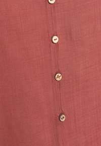 Chemise en lin rouge avec une finition texturée, dotée de six boutons ronds en bois le long de la patte de boutonnage et d'un design à coupe décontractée.