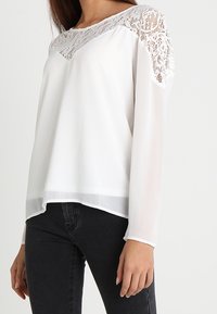 Top bianco a maniche lunghe con dettaglio in pizzo sulle spalle, caratterizzato da una vestibilità rilassata e una lieve trasparenza lungo l'orlo.