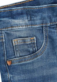 Kék denim anyag, textúrált felülettel. Kis elülső zseb és szív alakú fém dísz található rajta. Látható sárgásbarna varrás.