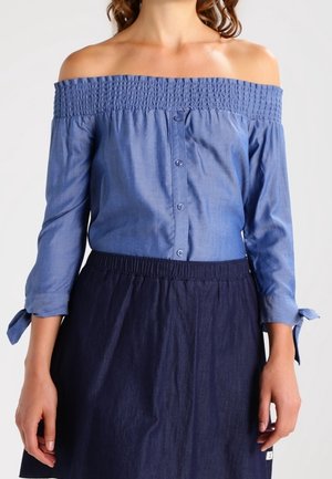 Nainen, jolla on yllään sininen olkapäät paljastava paita, jossa on napit edessä ja kolmen neljänneksen hihat, jotka on sidottu kalvosimista, yhdistettynä tummansiniseen hameeseen.