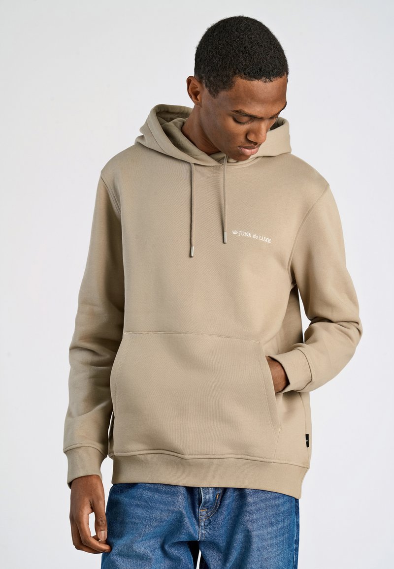 Felpa beige con cappuccio e cordino, tasca frontale e logo. Presenta un tessuto testurizzato e polsini a costine. Abbinata a jeans blu.