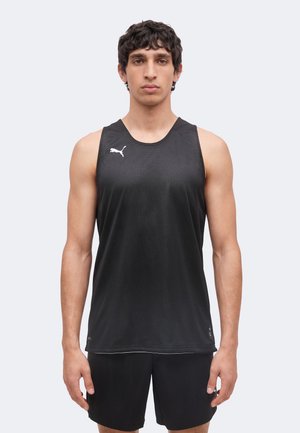 Jeune homme debout face à l'objectif, portant un débardeur de sport noir et un short noir sur un fond clair et uni.