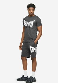 Jonge man die een zwart "TAPOUT" T-shirt en bijpassende korte broek draagt, witte sokken en zwarte sneakers, staand met één hand in zijn zak.