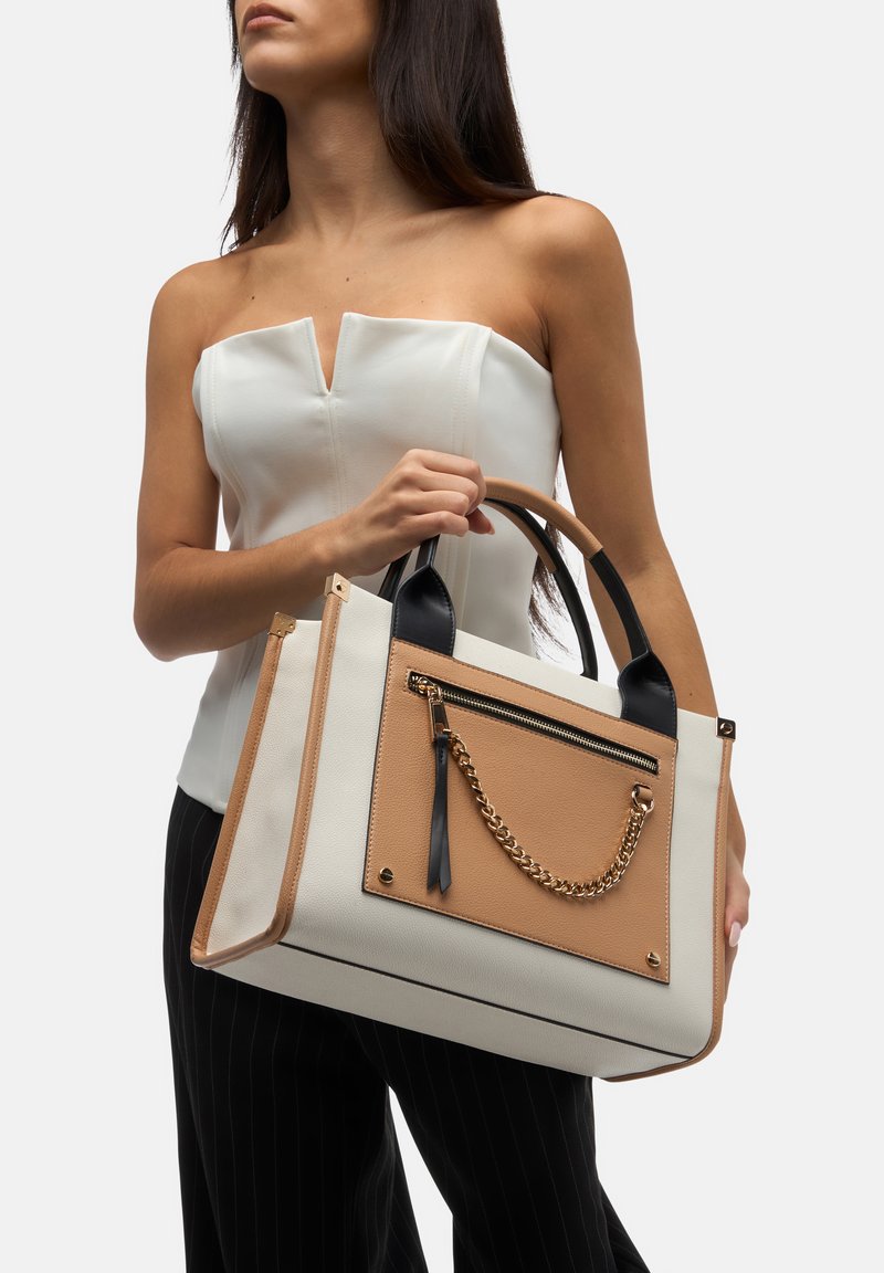 Sac fourre-tout en cuir synthétique crème et beige avec des poignées noires. Doté d'une poche zippée à l'avant avec un accent en chaîne dorée et des rivets.