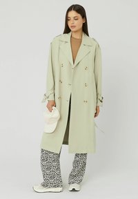Femme portant un long trench-coat beige à double boutonnage, un pantalon à imprimé animal, des baskets blanches, tenant une casquette de baseball blanche, regardant vers le bas sur le côté.