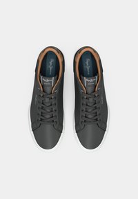 Ζευγάρι μαύρα χαμηλά sneakers με λευκές σόλες και καφέ επενδεδυμένα κολάρα, από πάνω σε άσπρο φόντο.