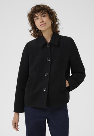 Femme aux cheveux bouclés portant un manteau noir boutonné avec des poches, debout devant un fond clair uni.