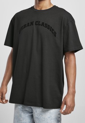 Czarny oversize'owy T-shirt z napisem "Urban Classics" w łukowatym, czarnym druku, noszony z jasnoniebieskimi dżinsami przez osobę o średnim odcieniu skóry.