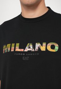 Črna bombažna majica s multi-barvnim grafičnim napisom "MILANO" in besedilom "PARCO LAMBRO" v krepki pisavi. Vključuje logotip EA7 Emporio Armani.