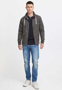 Tamno siva zip-up jakna s teksturiranim uzorkom, bijele vezice, uparena s crnom majicom i svijetloplavim trapericama. Nosena uz bijele tenisice.