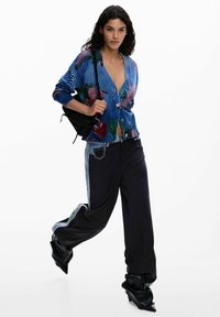 Blau-grün gemusterte Strickjacke mit Blumenmuster und Knopfverschluss, kombiniert mit schwarzen, weit geschnittenen Hosen mit Denim-Akzenten. Schwarze Tasche mit Herzanhänger.