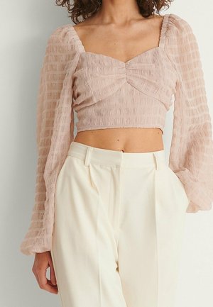 Frau trägt ein strukturiertes, hellrosa, langärmliges Crop-Top mit Puffärmeln und hochtaillierte, cremefarbene Hose, Hände in den Taschen.