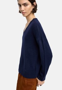 Granatowy sweter dzianinowy z dekoltem w serek, luźny krój, rękawy raglanowe i teksturowany materiał. W komplecie z brązowymi spodniami sztruksowymi.