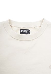 Hellbeige Sweatshirt mit geripptem Rundhalsausschnitt, ausgestattet mit einem schwarzen Etikett, auf dem "HONESTY RULES" steht. Weiche Textur, einfaches Design, minimalistische Details.