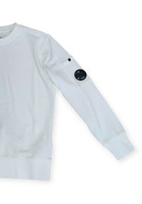 Felpa bianca con maniche lunghe, polsini a costine e una tasca a toppa sul braccio sinistro con un emblema circolare nero. Texture liscia.