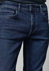 Tamsiai mėlynos džinsinės kelnės su klasikinio dizaino, penkiomis kišenėmis ir kontrastinga siuvinėta. Audinys turi švelnų tekstūrą. Standartinė liemens juosta su mygtuku.