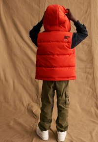 Gilet rembourré rouge avec capuche, logo au dos et manches noires, associé à un pantalon cargo vert et des chaussures blanches sur un fond neutre.
