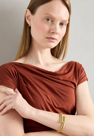 Top de tela drapeada en color óxido con mangas cortas. Modelo llevando una pulsera dorada con patrón en espiral en su muñeca. Fondo suave.