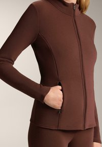 Smeđi zip-up jakna s dugim rukavima, visokim ovratnikom i bočnim džepovima. Glatka tekstura i prilagođeni dizajn poboljšavaju fit i funkcionalnost.