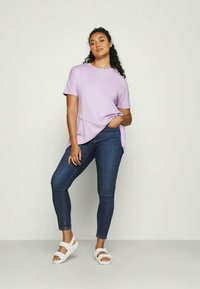 Svetlo purpurna majica kratkih rukava s zakrivljenim rubom, upairana s tamnoplavim uskim trapericama i bijelim sandalama. Glatka tekstura kroz cijelu površinu.