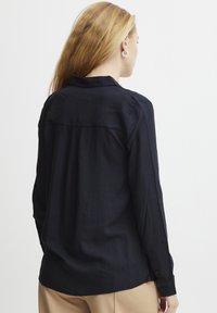 Maglietta a maniche lunghe blu navy con una consistenza morbida, colletto classico, dettaglio della cucitura posteriore e polsini con bottoni, abbinata a pantaloni beige.