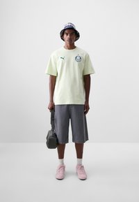 Camiseta de algodón verde claro con logo, pantalones cortos de mezclilla grises, zapatillas rosas y bolso de hombro gris. Sombrero tipo cubo con patrón de cuadros morados.